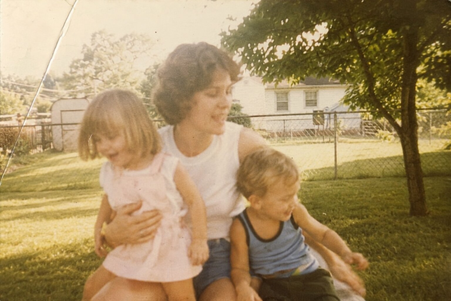 A faded 1980s family snapshot &mdash; a woman holding two young children in a backyard.
